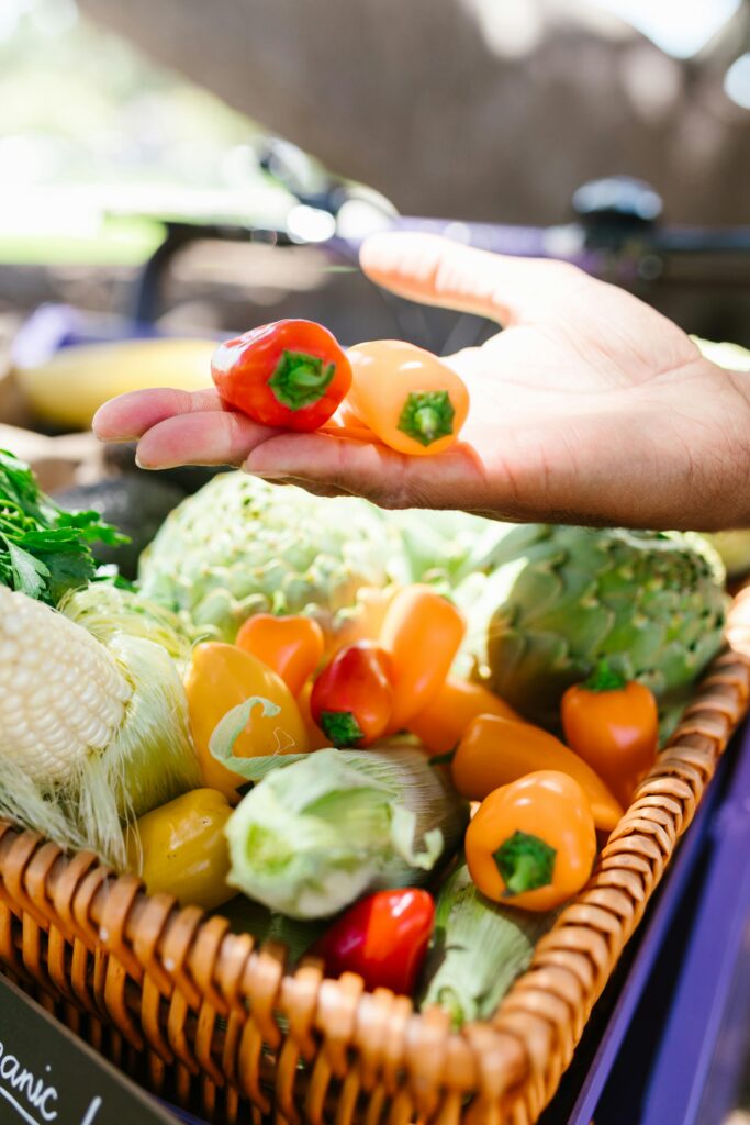 A hand picking fresh organic peppers from a basket filled with colorful vegetables at a farmers market.